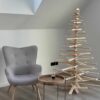 Tall Franky Holzweihnachtsbaum in minimalistischem Design, aufgestellt neben einem grauen Sessel und einem runden Holztisch, ideal für moderne Wohnräume.