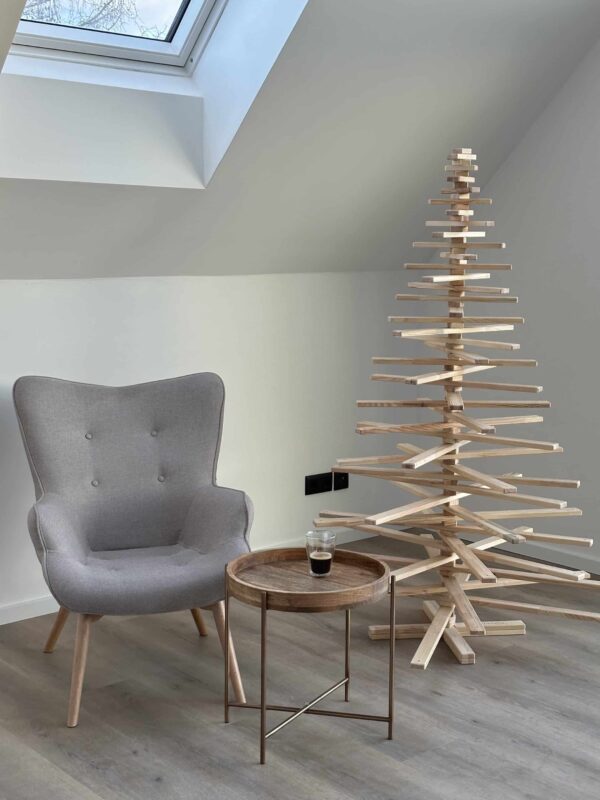 Tall Franky Holzweihnachtsbaum in minimalistischem Design, aufgestellt neben einem grauen Sessel und einem runden Holztisch, ideal für moderne Wohnräume.