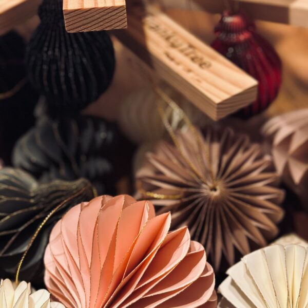 Detailaufnahme von nachhaltigen Papieranhängern in Wabenstruktur in verschiedenen Farben wie Rosa, Beige, Schwarz und Rot, dekorativ arrangiert unter einem Holzweihnachtsbaum.